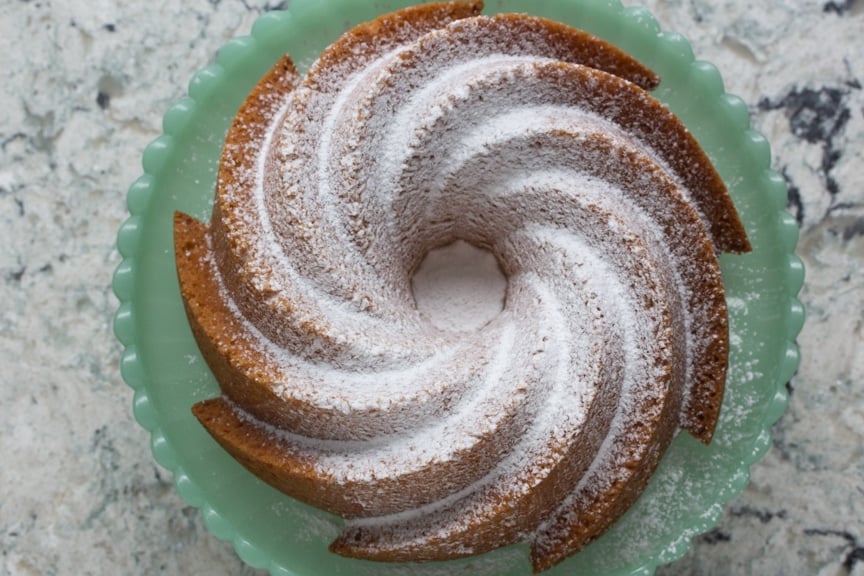 A beautiful almond butter cake in a swirl bundt pan pattern sprinkled with powdered sugar on a green milk glass plate. 