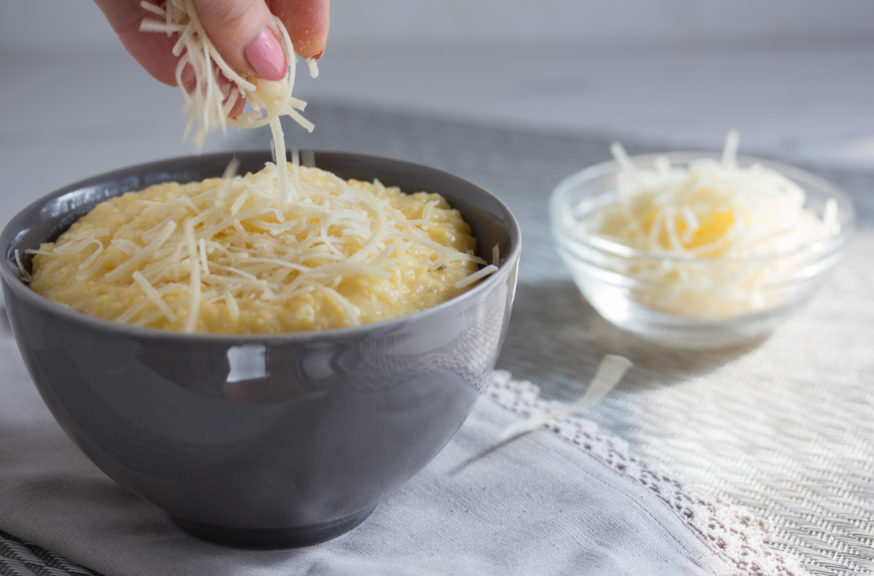 Shredded cheddar cheese being added to a bowl of creamy corn grits. 