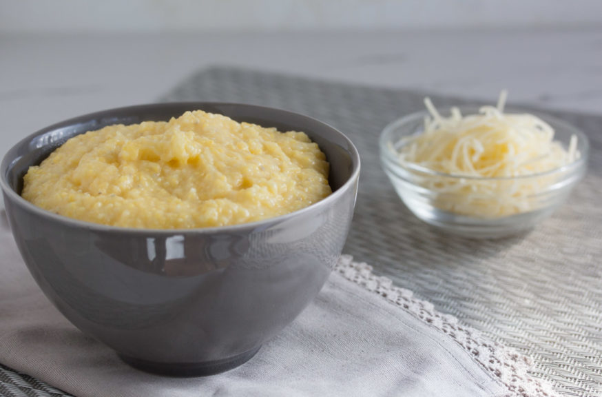 A bowl of creamy corn grits next to a bowl of shredded cheddar cheese. 