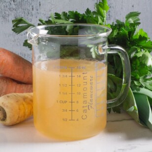 A glass measuring container filled with homemade Low FODMAP vegetable broth, in front of fresh carrots, parsnips, parsley and leeks.
