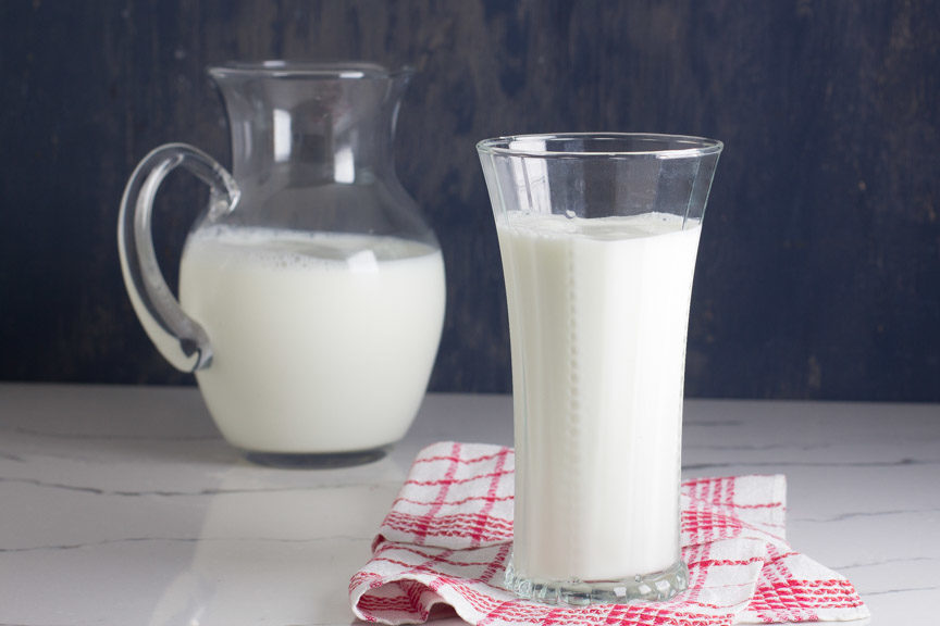 A tall glass of lactose free milk in front of a glass pitcher of milk. 