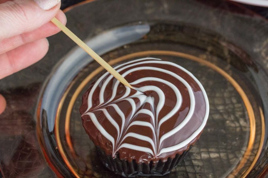 A hand holding a toothpick creating spider webs in ganache on top of a cupcake. 