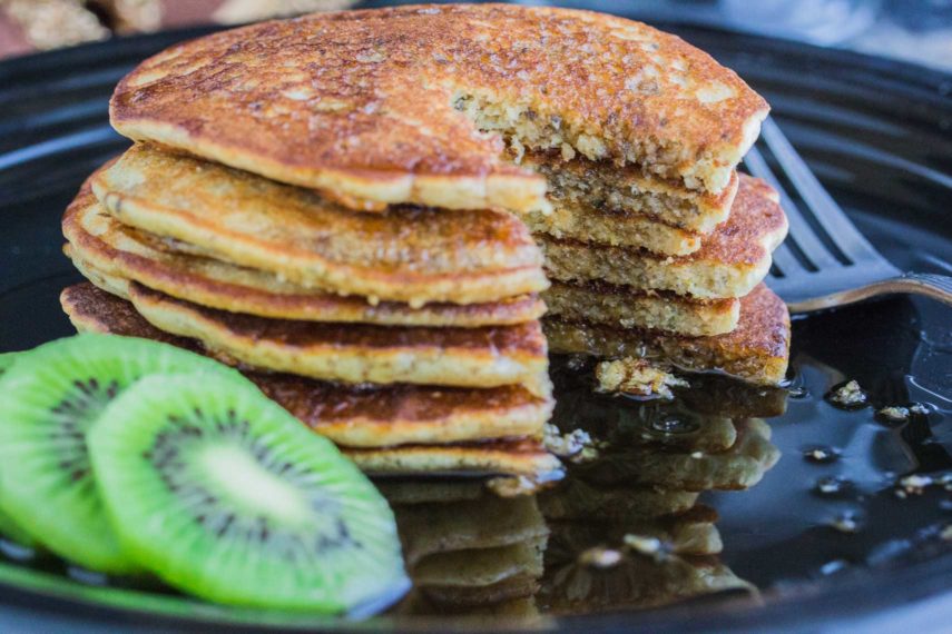 Maple syrup being poured over 3 Seed dairy-free gluten-free Pancakes on a plate served with sliced kiwi. With a slice taken out. 