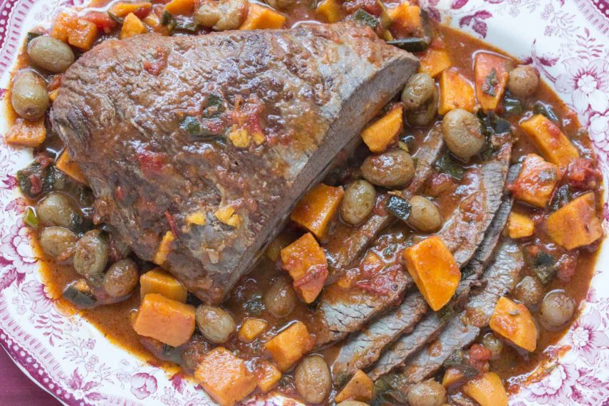 closeup overhead image of brisket with sweet potatoes & grapes in a red and white platter