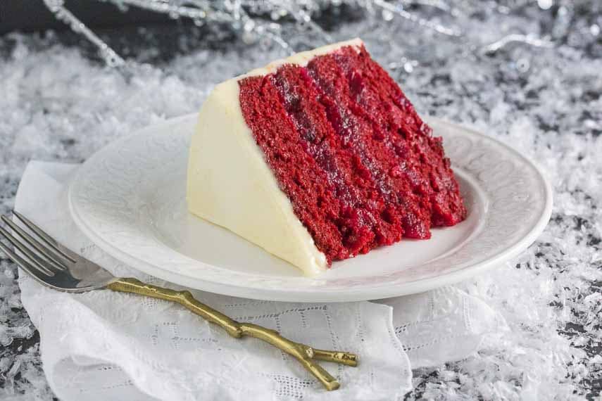 A slice of moist Red Velvet Cake with White Chocolate Frosting and Sugared Cranberries on a white plate. 