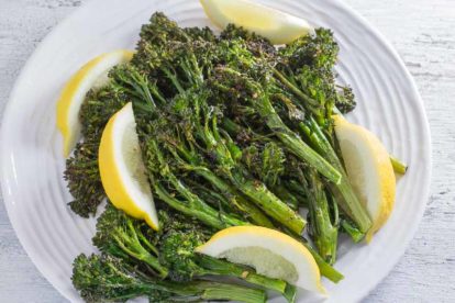 spicy roasted broccolini with garlic and lemon on a round white plate and a white wood background