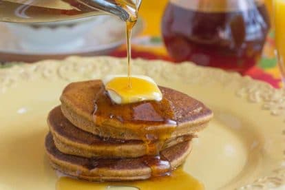 closeup of gingerbread pancakes on a yellow plate, butter melting on top and maple syrup being poured over the stack