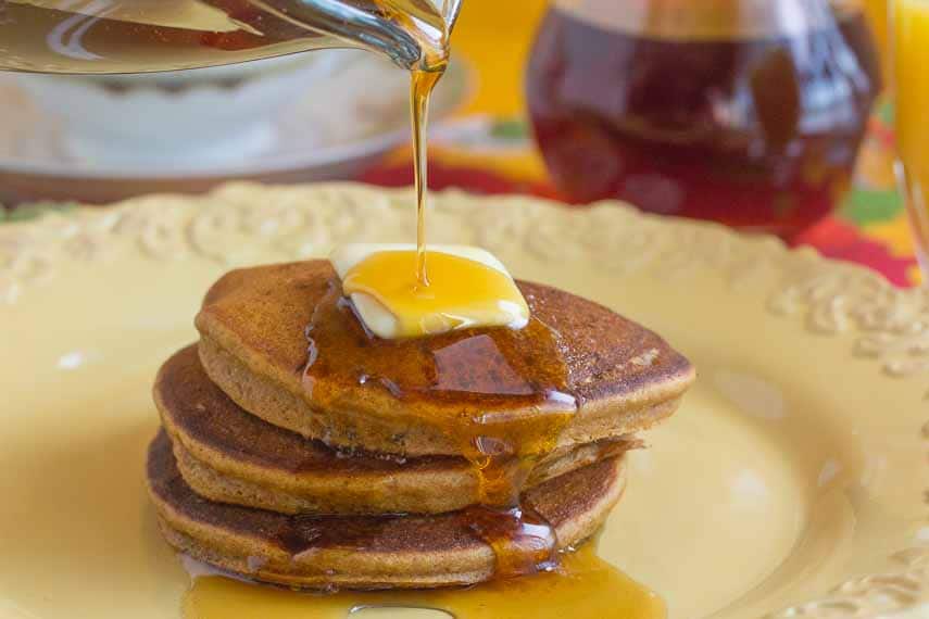 closeup of gingerbread pancakes on a yellow plate, butter melting on top and maple syrup being poured over the stack
