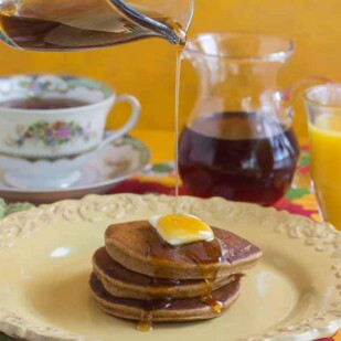 gingerbread pancakes on a yellow plate, butter melting on top and maple syrup being poured over the stack. Glass of orange juice in the background and a cup of tea.