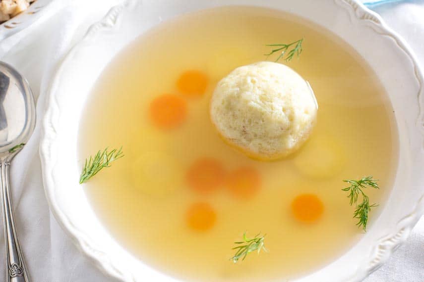 matzo ball soup in white bowl with fresh dill floating on top; silver spoon alongside