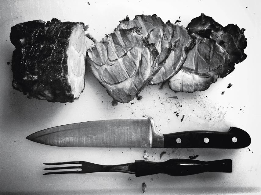 overhead black and white shot of sliced ham on a board with a carving knife and fork