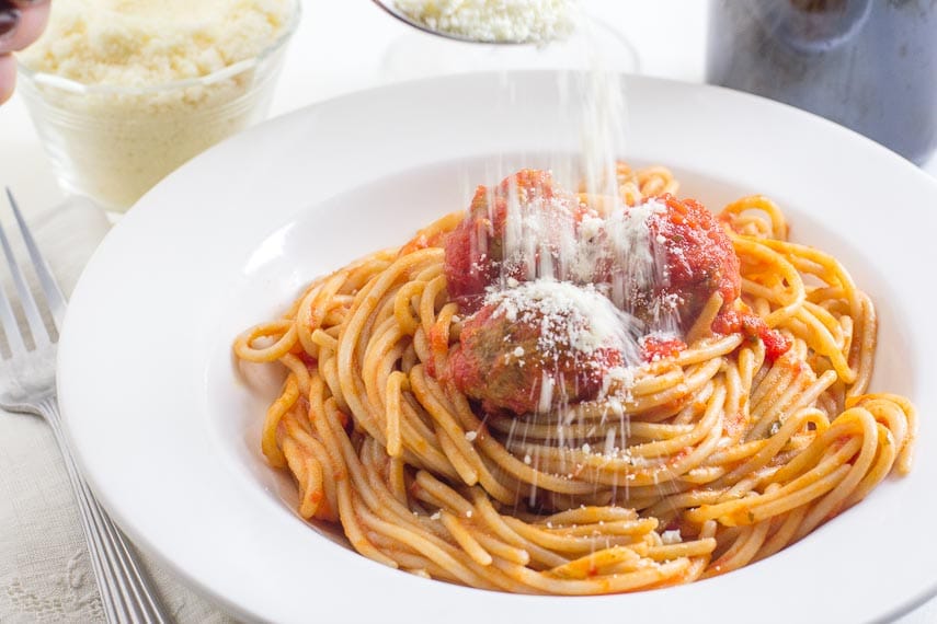 sprinkling Parmesan cheese over spaghetti & meatballs in a white bowl