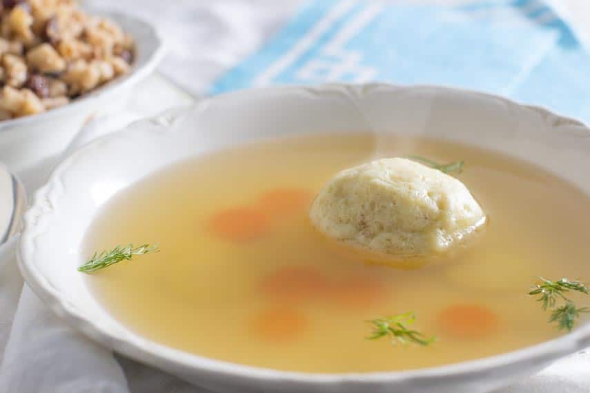 steam coming from a bowl of hot matzo ball soup in a white bowl; charoset in background