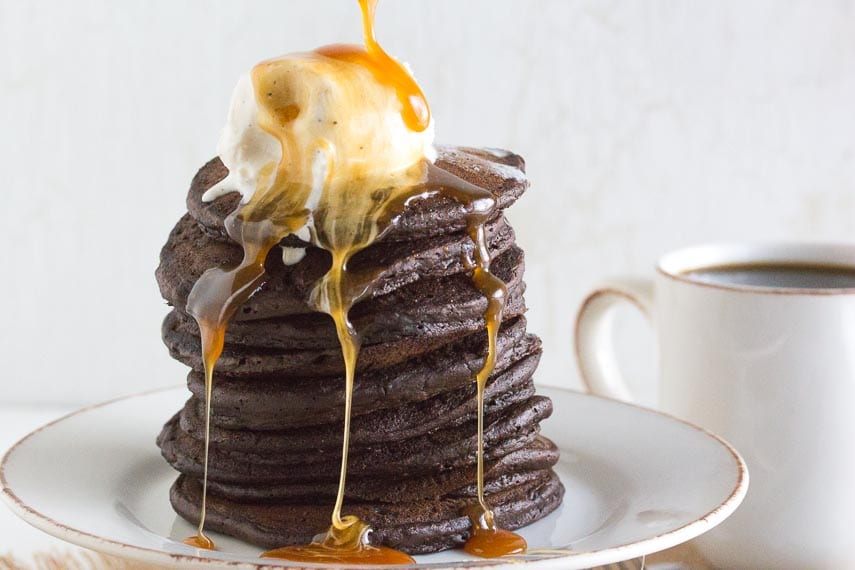 tall stack of chocolate pancakes with melting vanilla ice cream on top and salted caramel sauce dripping over sides