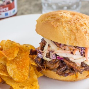 BBQ brisket topped with slaw on a roll with BBQ potato chips on a white plate