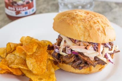 BBQ brisket topped with slaw on a roll with BBQ potato chips on a white plate