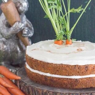 low FODMAP carrot cake with cream cheese frosting and bare sides on a wooden plate with decorative bunny in the background