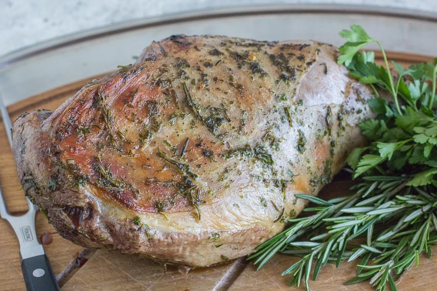 roast leg of lamb on a cutting board with fresh herbs alongside