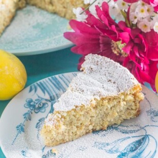 slice of lemon almond cake on a blue and white platye; pink and white flowers in background along with lemons