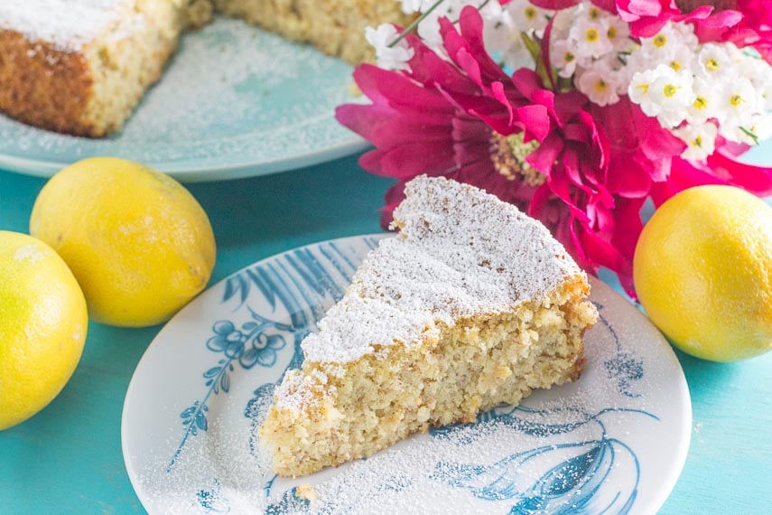 slice of lemon almond cake on a blue and white plate; pink and white flowers in background along with lemons