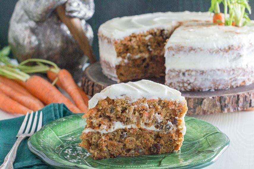 slice of low FODMAP carrot cake with cream cheese frosting on green plate with carrots and whole cake in background