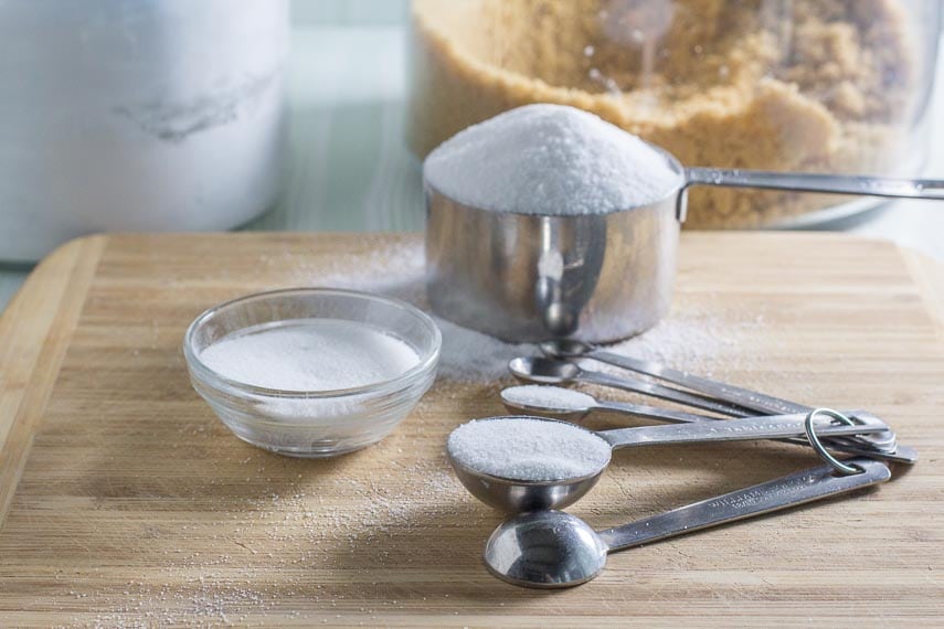 All About Sugar. White sugar in measuring cups, spoons and a bowl on a wooden board. Brown sugar and confectioners' sugar in background.