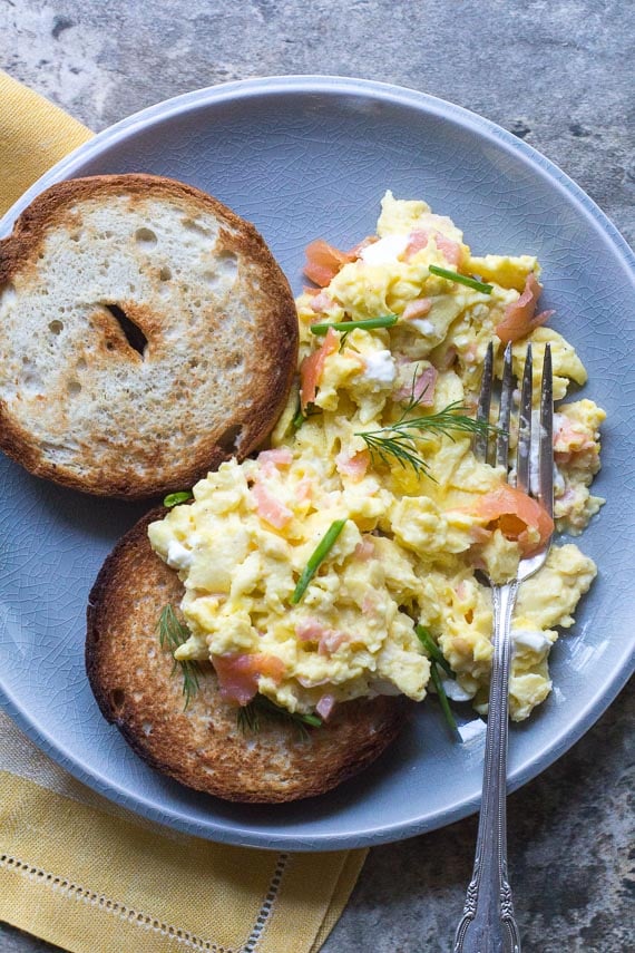 vertical image of scrambled eggs with smoked salmon, cream cheese, chives nad dill on a crackled plate with toasted bagels