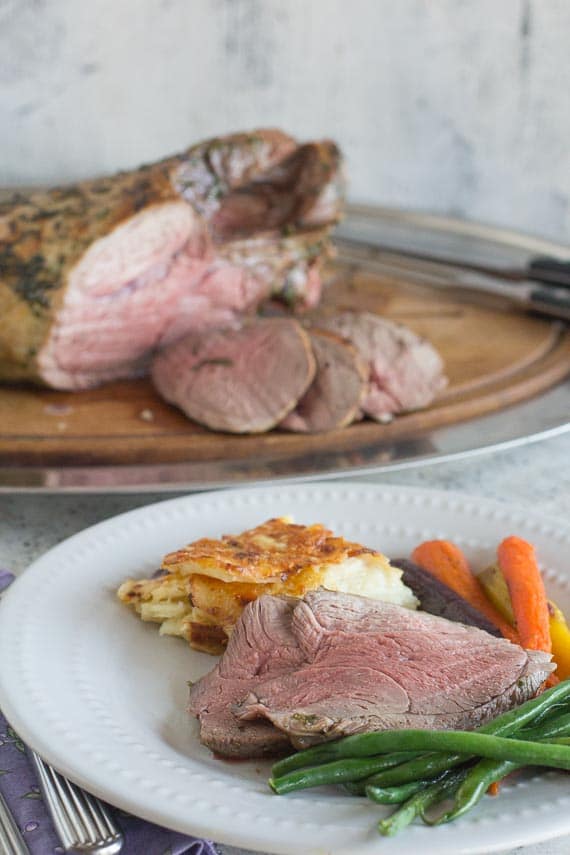 vertical image of sliced leg of lamb on a plate with roasted carrots and steamed green beans