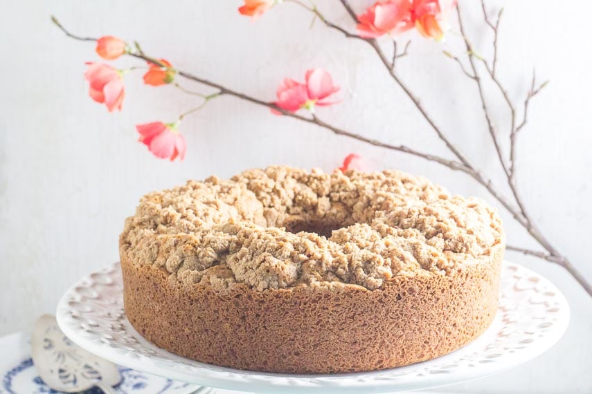 unadorned cinnamon streusel coffee cake on white platter with red blossoms in the background