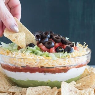 hand holding chip digging into 7 layer dip in glass bowl