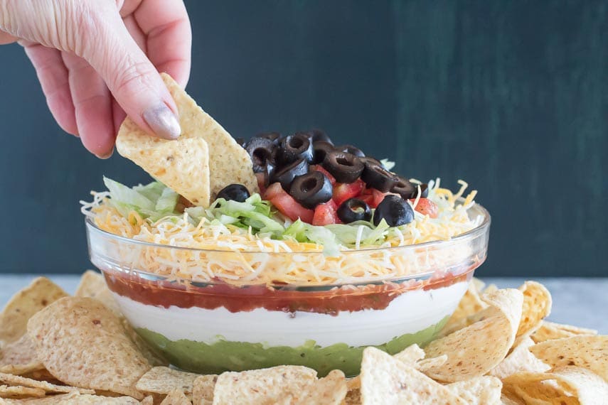 hand holding chip digging into 7 layer dip in glass bowl
