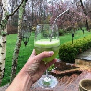 hand holding wine glass with Cucumber Mint Lime Agua Fresca with straw against garden backdrop