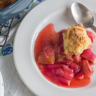 overhead image of serving of strawberry rhubarb cobbler on white plate on top of floral linens
