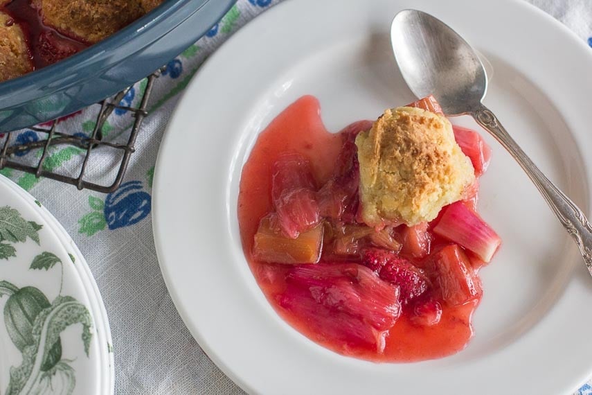 overhead image of serving of strawberry rhubarb cobbler on white plate on top of floral linens