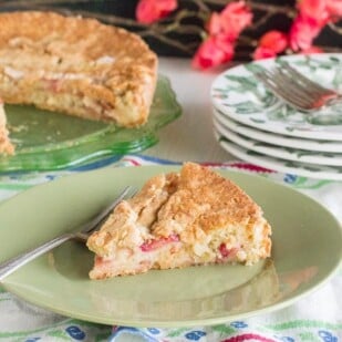 rustic rhubarb cake slice on green plate, whole cake in background on green glass plate. Floral linens and red blossoms in background
