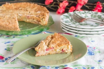 rustic rhubarb cake slice on green plate, whole cake in background on green glass plate. Floral linens and red blossoms in background