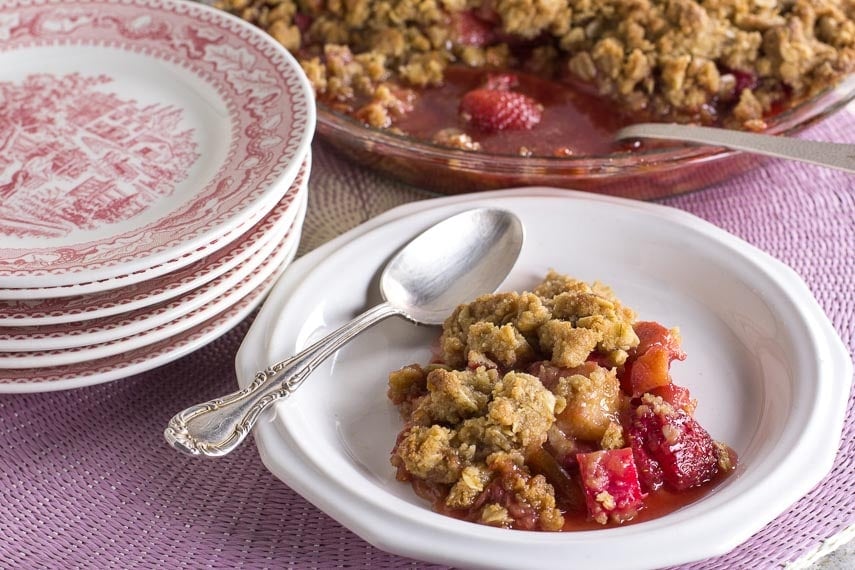 strawberry rhubarb crisp on a white plate with silver spoon; stack of pink plates and whole crisp in background