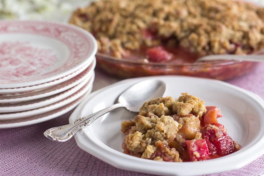 strawberry rhubarb crisp on a white plate with spoon alongside; stack of pink plates and whole crisp in background