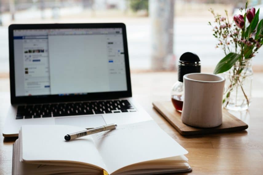 Desk layout with an open laptop and notebook with a pen and cup of coffee.