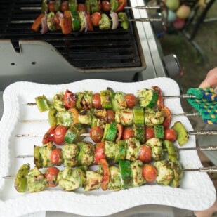 placing grilled chicken and vegetable kebobs on a white platter