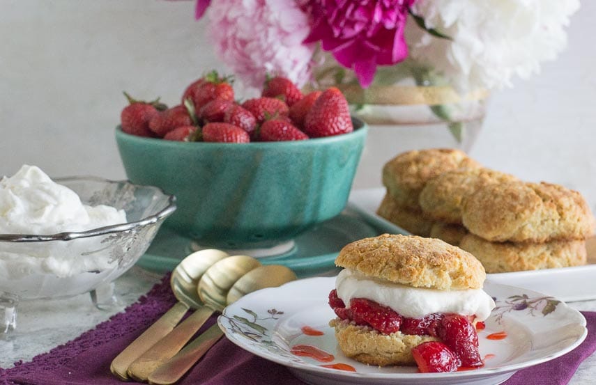 strawberry shortcake on a white plate with gold spoons alongside; berries and peonies in background