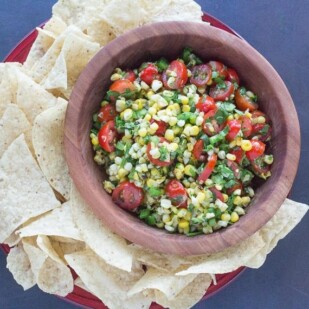 overhead of grilled corn salsa in wooden bowl on red plate with side of corn chips