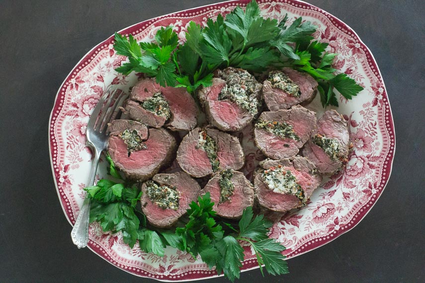 Beef Tenderloin Stuffed with Goat Cheese, Spinach & Sun-Dried Tomatoes on red and white platter with fresh parsley