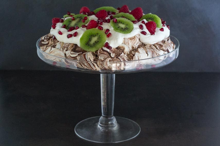side view of Low FODMAP Chocolate Pavlova with Pomegranate, Raspberries & Kiwi in a glass pedestal dish