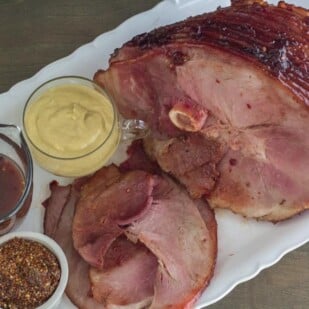 Low FODMAP Cranberry Hot Pepper Glazed Ham on oval white platter with pan juices and assorted mustard