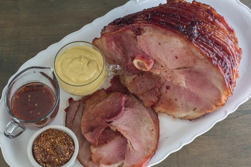 Low FODMAP Cranberry Hot Pepper Glazed Ham on oval white platter with pan juices and assorted mustard