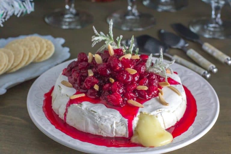A round wheel of brie topped with a rich cranberry sauce, slivered almonds, and sprigs of rosemary on a white plate. Melted cheese oozes from the side. Crackers and utensils are in the background.
