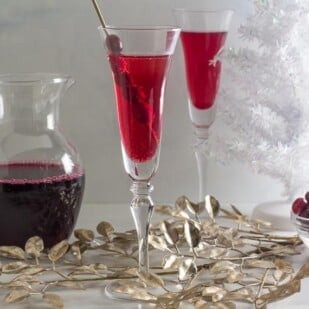 Low FODMAP Cranberry Cinnamon Swizzle in a champagne glass; cranberry juice in pitcher