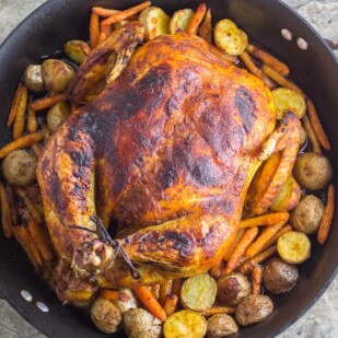 overhead image of whole roasted curry chicken with potatoes and carrots in a roasting pan against a stone backdrop