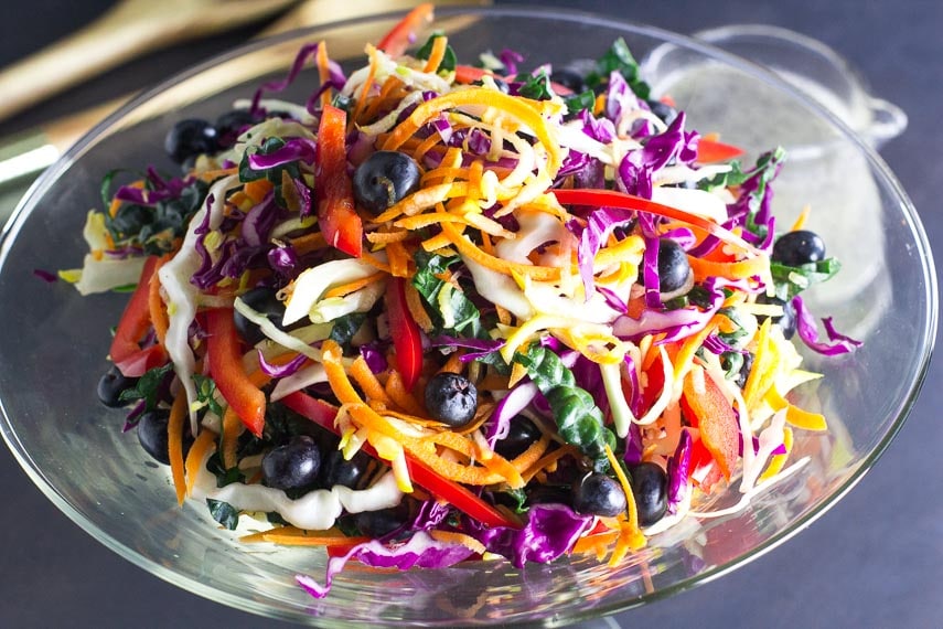 closeup of Low FODMAP Eat the Rainbow Slaw in a clear glass bowl; dressing and serving implements in background